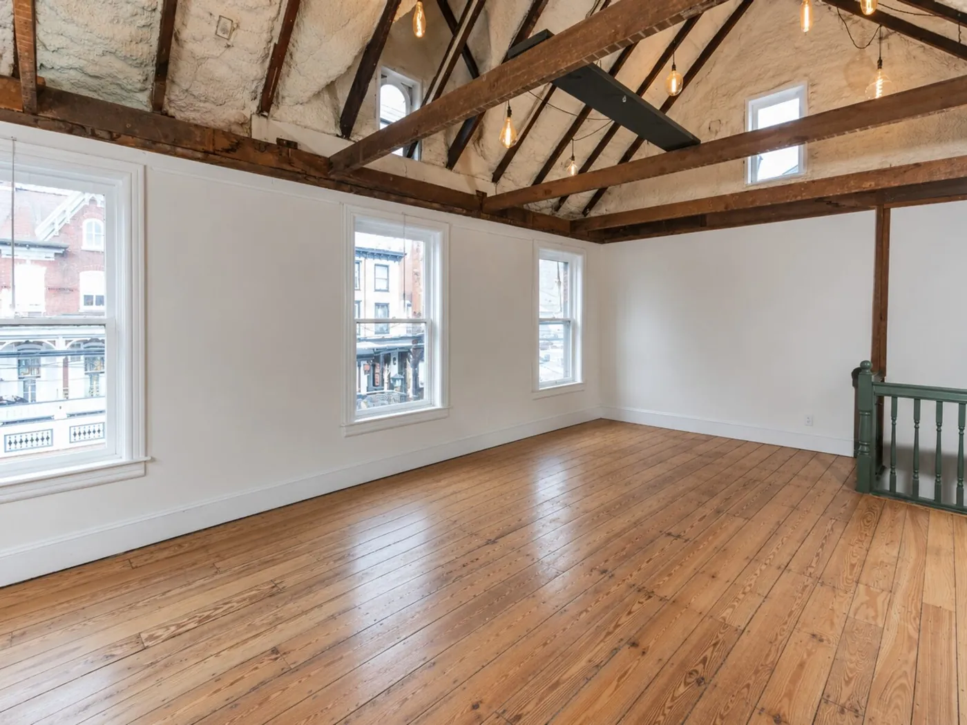 An upstairs room with exposed timber rafters, string lights, tall windows and wide pine floorboards